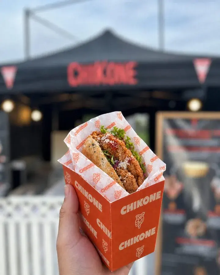 Eine Hand hält das Baguette di Parma mit Chicken Tenders, Parmesan, Soße und Salat in der Chikone-Box, dahinter der Chikone-Stand