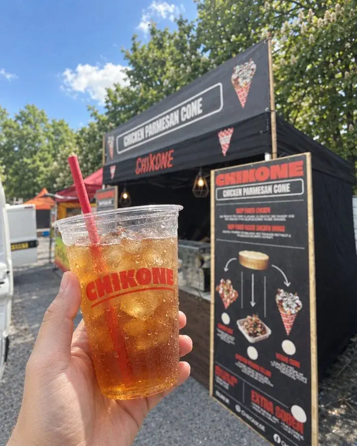 Eine Hand hält einen frischen Eistee im Becher mit Chikone-Logo, dahinter der Chikone-Stand mit Banner und Speisekarte